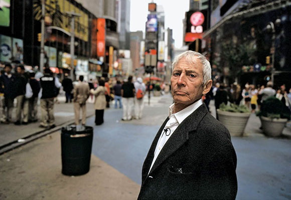 An older man with white hair in a dark blazer looks toward the camera in a crowded city square.