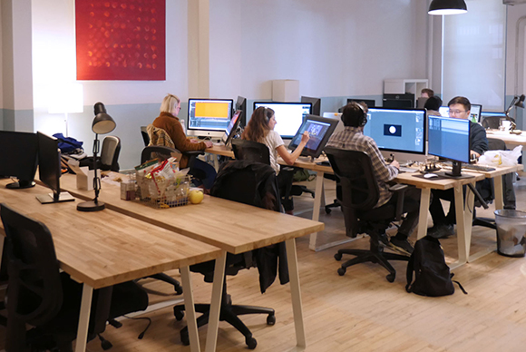 Several people sit at long wooden desks using desktop computers in a bright, open office environment.