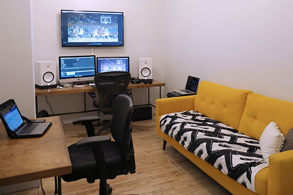A workspace with two desks, multiple computer monitors, speakers, and a yellow sofa in a room with light walls.