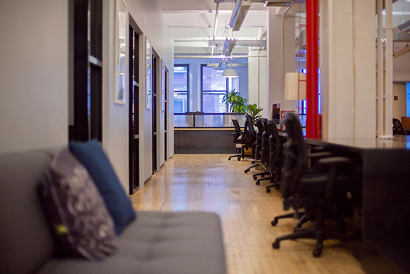 A long office desk with black chairs lined up in a modern, brightly lit workspace with wooden floors.