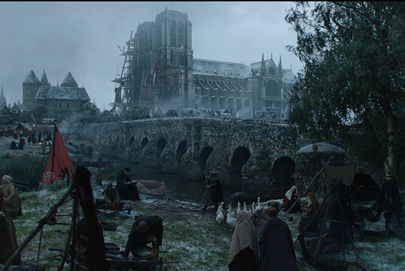 A large stone castle with scaffolding and a stone bridge over a river near a small village.