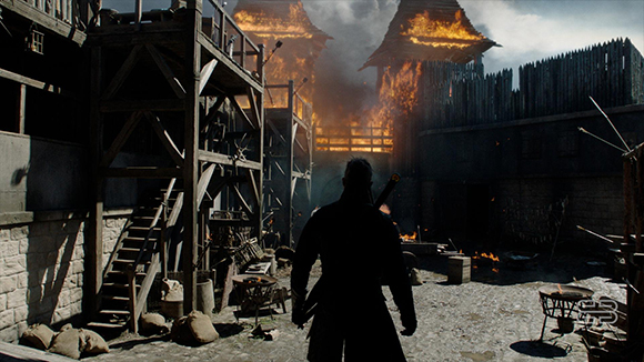 Silhouette of a man standing in a courtyard facing two wooden towers engulfed in flames.