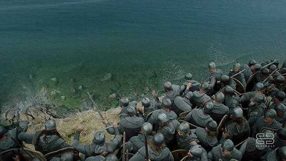 A crowd of soldiers in chainmail and helmets standing on a rocky coastline facing the ocean.