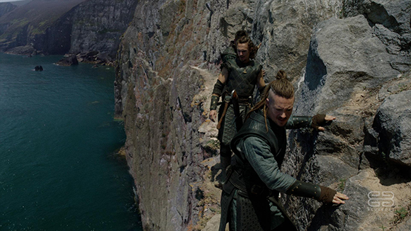 Two people in dark, textured clothing climb a steep, rocky cliff overlooking a deep blue sea.