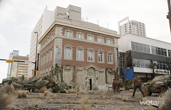 A two-story brick building with arched windows sits surrounded by rubble and dry weeds in a city.