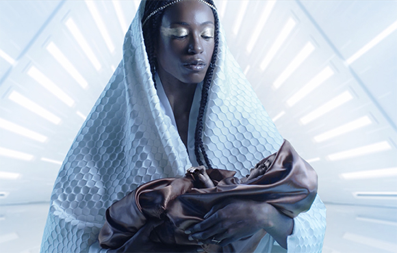 A person with gold eye makeup wearing white textured robes holds a baby wrapped in brown cloth.