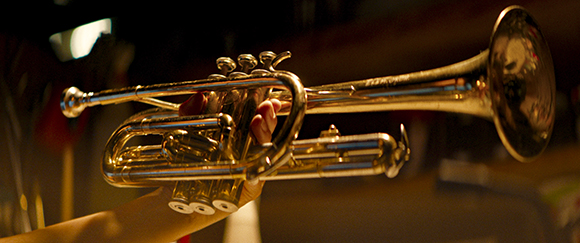 A person's hands holding a shiny brass trumpet against a dark, blurred background.