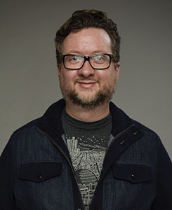 A man with brown hair, glasses, and a beard wearing a dark denim jacket over a graphic t-shirt.