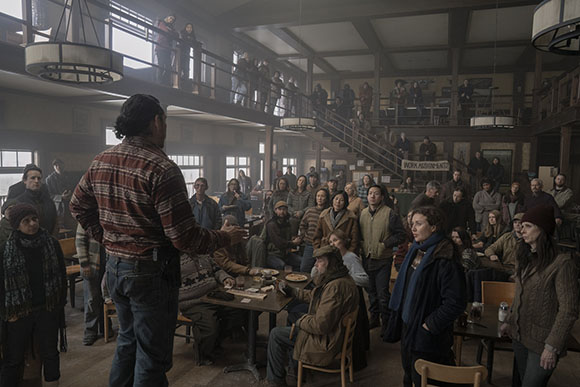 A group of people sit and stand in a dimly lit, rustic room with wooden beams and stairs.