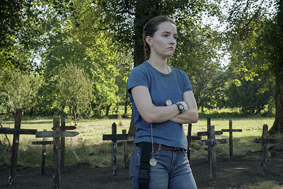 A young woman with braided hair stands with arms crossed in a field of wooden crosses.