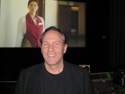 A smiling man in a black jacket stands before a screen displaying a woman in a red uniform vest.