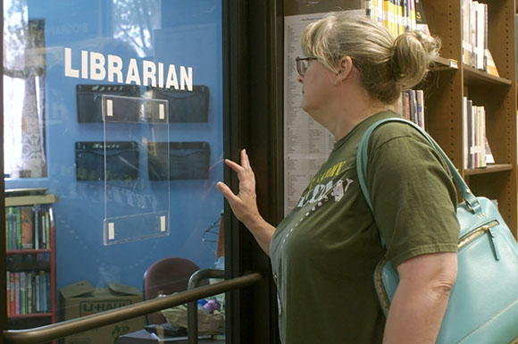 A woman with grey hair in a bun looks through a glass door labeled LIBRARIAN.