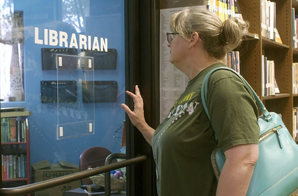 A woman with blonde hair in a bun looks through a glass door with the word Librarian printed on it.