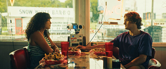 A young woman with curly hair and a young man sit across from each other at a diner table.