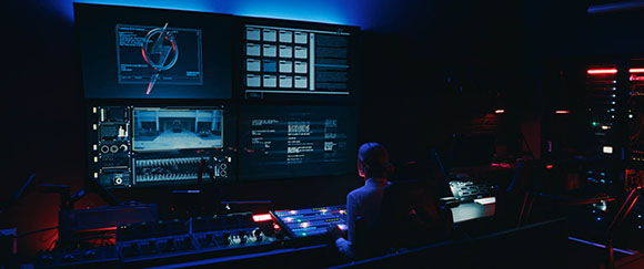 A person sits in a dark room facing a large wall of glowing digital monitors and control panels.