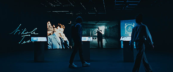 Silhouetted people walk past large, glowing museum displays in a dark room.