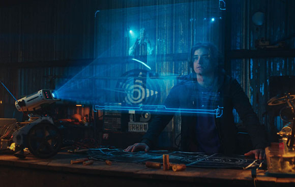 A person sits at a workbench looking at a blue holographic display projected from a small device.