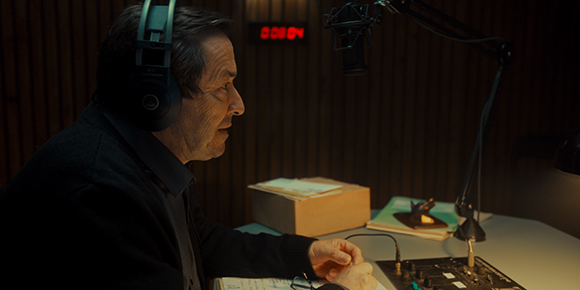 An older man with headphones sits at a desk with a microphone and audio equipment in a dark studio.