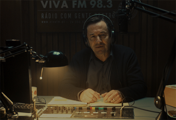 A man wearing headphones sits at a desk with a microphone and mixing console in a radio studio.