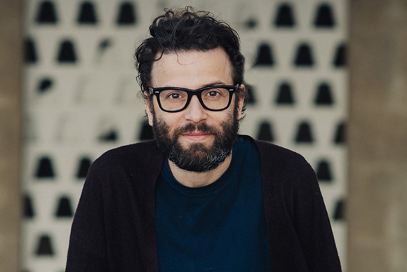 A man with dark wavy hair, a beard, and black-rimmed glasses looking at the camera.
