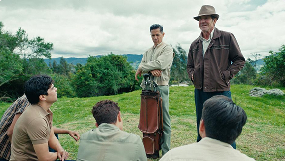 Two men stand while three others sit in a grassy field against a backdrop of green hills.
