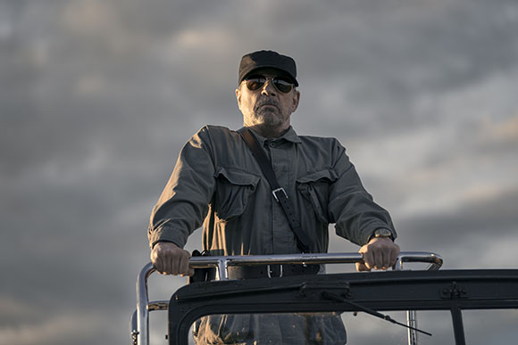 A man wearing a black cap, sunglasses, and a grey utility shirt stands behind a metal vehicle railing.