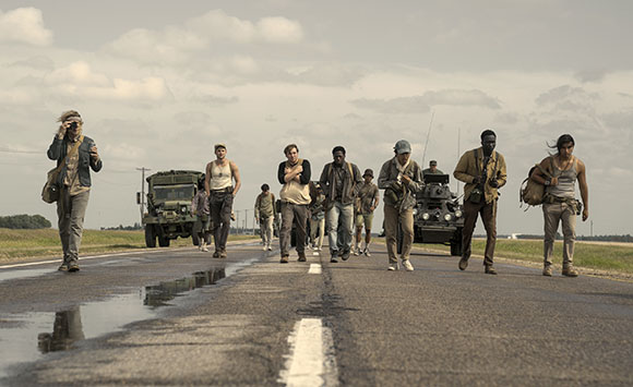 A group of people walking along a paved road with military vehicles in the background.