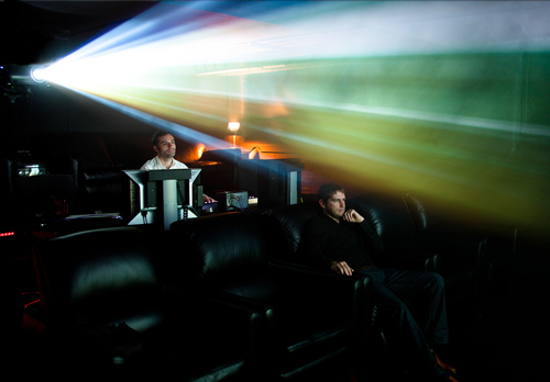 A bright, multicolored light beam from a projector cuts through a dark theater where two men are seated.
