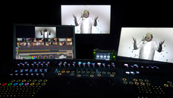 A dark editing studio featuring three monitors displaying video footage and a large control panel with glowing buttons.