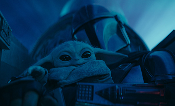 A small green creature with large eyes looks up toward a dark metallic helmet.