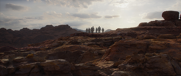 A group of figures walking across a vast, rocky desert landscape under a bright, cloudy sky.