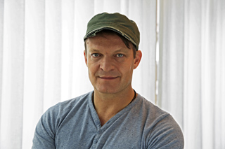 A man in a green cap and grey v-neck shirt looking at the camera against a white curtain background.