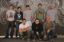 Eight diverse people posing together in two rows against a black and white swirling pattern wall.