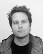 Black and white headshot of a man with messy hair wearing a jacket and dark shirt.