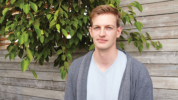 A man with light brown hair wearing a white v-neck shirt and a grey cardigan stands before a wooden wall.