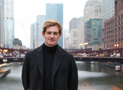 A man with blond hair wearing a black turtleneck and coat, standing before a city river and skyscrapers.