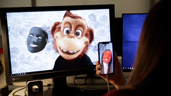 A person holds a smartphone displaying a red mask in front of a monitor showing a cartoon monkey.