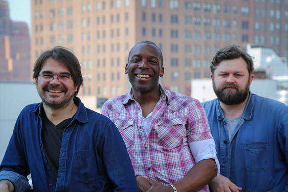 Three men smiling while standing together outdoors with a large brick building in the background.