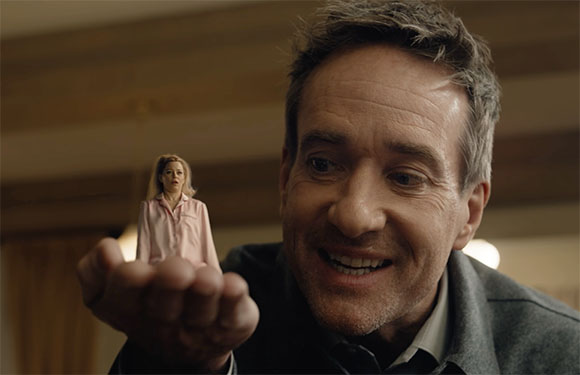 A man smiles as he holds a tiny, miniature woman in the palm of his hand.