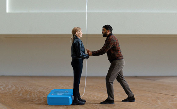 A man in a plaid shirt reaches toward a woman in a leather jacket standing near a white rope.