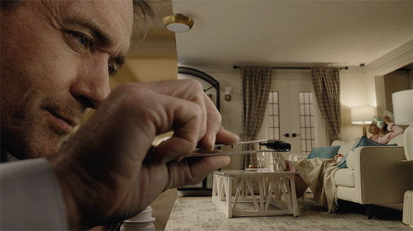 A man's hands hold a thin object in the foreground, with a person resting in a chair in the background.