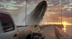 A large whale breaches from the water next to a bridge at sunset near a white pickup truck.