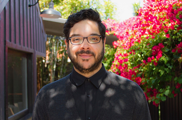 A man with dark hair, glasses, and a beard wearing a black shirt and grey cardigan, smiling in front of pink flowers.