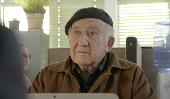 An elderly man with a dark cap and brown jacket looks ahead in an office setting.