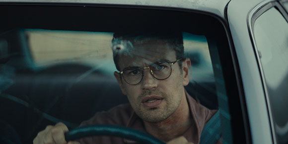 A man wearing glasses sits in the driver's seat of a car behind a cracked windshield.
