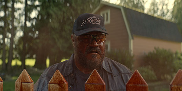 A man with a beard and sunglasses wearing a black El Rancho Motel cap in front of a house.