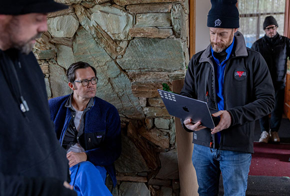 A man in a black jacket holds a laptop while looking at another man sitting against a stone wall.