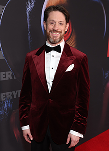 A man with a beard wearing a burgundy velvet tuxedo jacket and black bowtie.