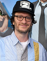 A man with glasses and a black baseball cap smiling in a light blue button-down shirt.