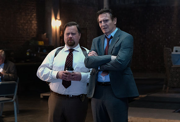 A man in a white shirt and red tie stands next to a man in a grey suit with arms crossed.